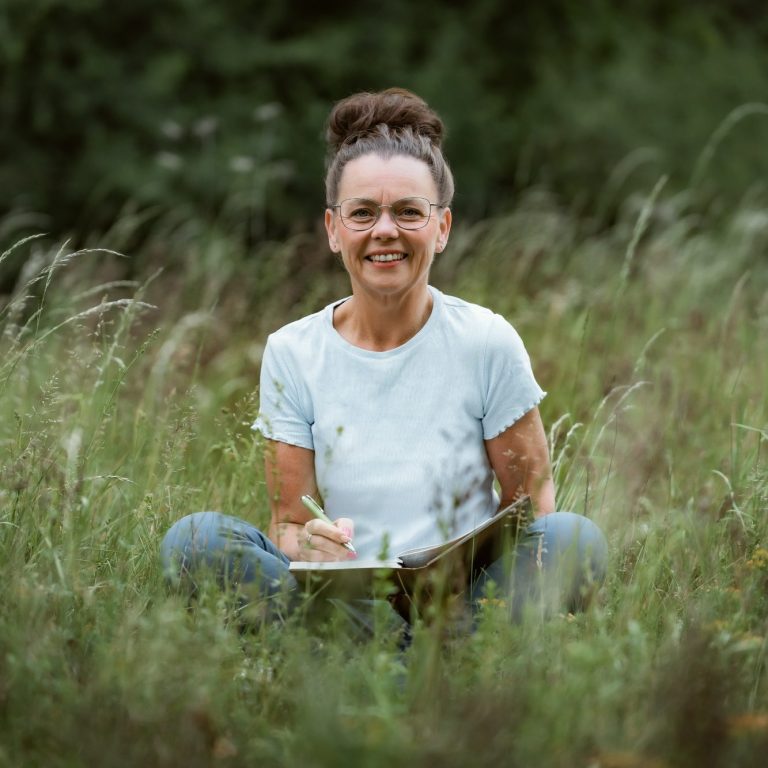 Traurednerin Kati Kreyser sitzt im Gras, lächelnd und schreibt in einem Notizbuch eure Hochzeitsrede.