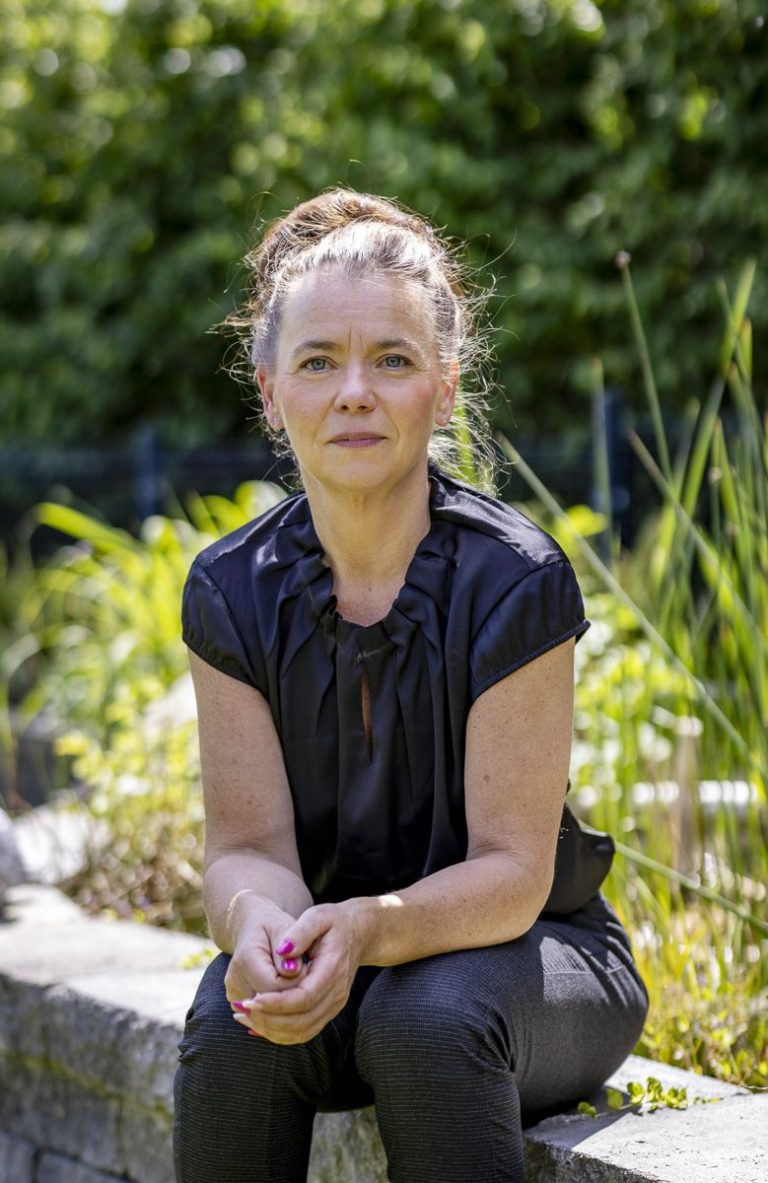Trauerrednerin Kati Kreyser mit schwarzer Bluse sitzt im Freien, umgeben von Pflanzen und grüner Landschaft.